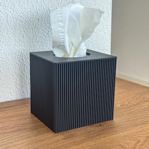 Puede incluir: Un dispensador de pañuelos negro con un diseño de costillas verticales. Los pañuelos blancos sobresalen de la abertura superior. El dispensador está sobre una superficie de madera. El texto "Nuvia: Onyx" está en la esquina inferior derecha.