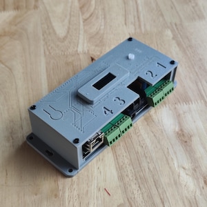 Op de afbeelding: Een grijs elektronisch apparaat met groene connectoren en zwarte schroeven. Het apparaat heeft de nummers 1, 2, 3 en 4 erop gedrukt. Het heeft ook een Ethernet-poort en een kleine rechthoekige opening. Het apparaat ligt op een houten oppervlak.
