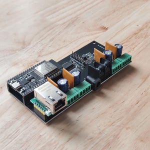 Op de afbeelding: Een zwart circuitboard met diverse elektronische componenten, waaronder een netwerkpoort, condensatoren en groene klemmenblokken. Het bord heeft een rechthoekige vorm en ligt op een houten oppervlak.