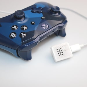 Könnte beinhalten: Ein blau-marinefarbener Videospiel-Controller mit Tarnmuster, einem weißen Kabel und einem weißen Stecker. Der Controller hat schwarze Tasten und ein schwarzes Steuerkreuz. Das Xbox-Logo ist sichtbar.
