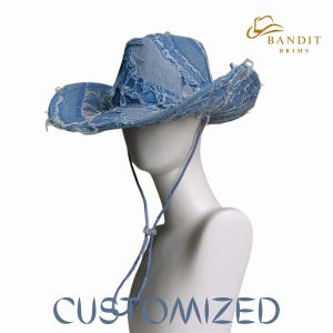 May include: A light blue denim cowboy hat with a distressed, frayed design. The hat features a wide brim and a chin strap. The word "CUSTOMIZED" is printed in blue at the bottom of the image. The hat is displayed on a white mannequin head.