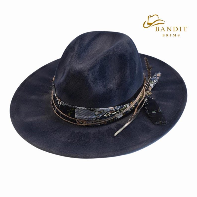 May include: A navy blue fedora hat with a wide brim. The hat features a decorative band with a floral pattern, rope detailing, and a feather accent. The hat has the text "BANDIT BRIMS" in gold.