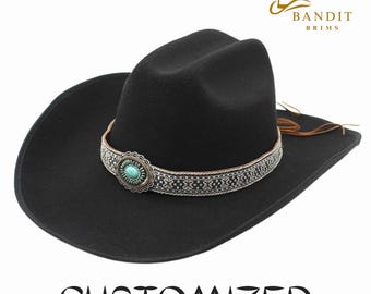 Pode incluir: Chapéu de cowboy preto com uma faixa decorativa com uma pedra turquesa e detalhes prateados. O chapéu tem uma aba larga e uma fivela de couro. A palavra "CUSTOMIZED" está impressa na parte inferior.