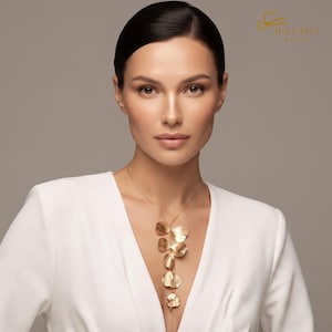 May include: A gold-toned necklace featuring a vertical arrangement of five overlapping, petal-shaped metal elements suspended from a delicate chain. The jewelry is worn over a white garment. The text "BANDIT BRUGS" appears in the top right corner.