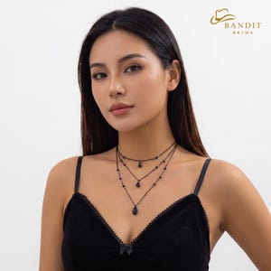 May include: A three-layered black necklace featuring delicate chains with small black beads and three dangling teardrop-shaped black pendants. The necklace is worn with a black velvet camisole featuring a small bow at the center of the neckline.