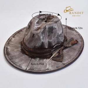 May include: A distressed brown hat with white accents, a dark brown band, and feather embellishments. The hat's crown measures 22.02" to 22.81" (56-58cm) across, with a brim width of 3.15" (8cm) and a height of 4.72" (12cm). The hat is from Bandit Brims.