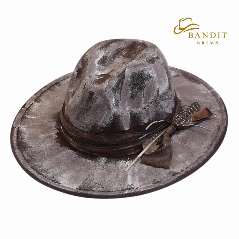 May include: A distressed brown fedora hat with white paint strokes. The hat features a brown leather band and is adorned with feathers. The hat has a wide brim and the logo "BANDIT BRIMS" is visible.