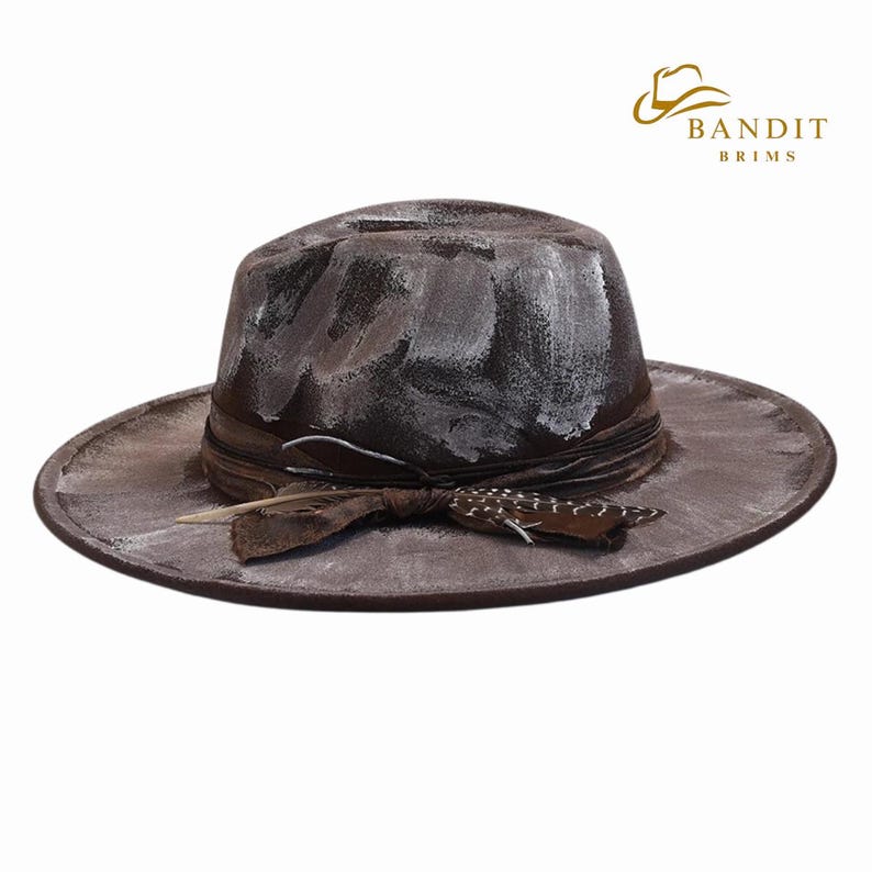 May include: A distressed brown fedora-style hat with white paint strokes. The hat features a brown ribbon band with a feather and decorative accent. The hat has a wide brim and the brand name "BANDIT BRIMS" is visible.
