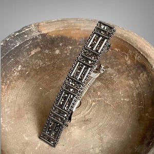 May include: A silver-toned hair clip with a rectangular design. The clip is adorned with small, dark-colored stones arranged in a geometric pattern. The clip is resting on a textured, earthy-toned surface.