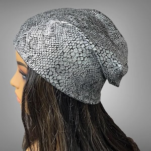 May include: A slouchy beanie hat featuring a metallic silver finish with a textured, black and silver snakeskin-inspired pattern. The fabric has a reflective quality, creating a shimmering appearance across the surface of the headwear.