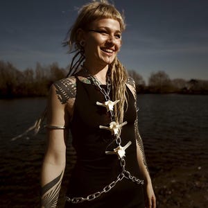 Pode incluir: Uma mulher vestindo um vestido preto e um colar de corrente de prata com pingentes de osso. A mulher tem tatuagens nos braços e ombros. Ela está sorrindo perto de um corpo de água. Joias únicas.