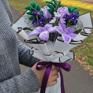 Puede incluir: Un ramo de flores moradas y blancas hechas a mano, envuelto en papel blanco con un diseño floral negro. El ramo está atado con una cinta morada. Las flores incluyen clemátides, tulipanes y otras variedades florales.