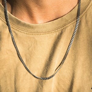 Könnte beinhalten: Eine silberne Halskette mit flachem Curb-Link-Design. Die Halskette wird über einem beigefarbenen T-Shirt getragen. Die Kette ist ein einfacher, klassischer Stil, der für den Alltag geeignet ist. Die Halskette ist ein beliebtes Accessoire.