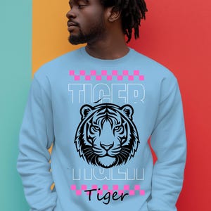 May include: Light blue sweatshirt with a black and white tiger face graphic. The word "TIGER" is printed above the tiger, and the word "Tiger" is printed below the tiger. Pink and white checkered patterns are above and below the word "TIGER".