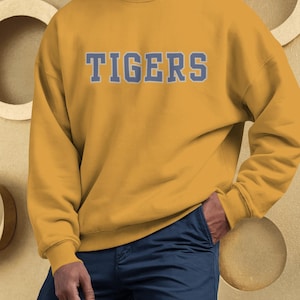 Op de afbeelding: Mosterdgeel sweatshirt met het woord "TIGERS" in lichtblauwe blokletters over de borst. De sweatshirt heeft lange mouwen en een ronde hals. De persoon op de foto draagt een marineblauwe broek.