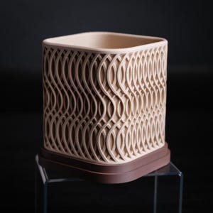 Peut inclure: Un récipient décoratif beige de forme carrée avec une base marron. L'extérieur du récipient présente un motif ondulé répétitif. Le design est ajouré, laissant passer la lumière. Probablement pour la décoration intérieure.