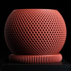 Può includere: Un oggetto decorativo sferico color terracotta con un motivo a reticolo. L'oggetto poggia su una base abbinata con creste verticali. Il design è simmetrico e la forma generale è rotonda.
