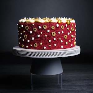 Puede incluir: Un pastel de terciopelo rojo decorado con chispas blancas y doradas sobre un soporte para pastel blanco.