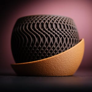 May include: A dark brown, spherical decorative object with a wave-like, openwork design. It sits atop a curved, tan-coloured base. The background is a gradient of dark and light pink.