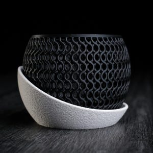 Peut inclure: Un pot de fleur sphérique noir avec un motif ajouré répétitif. Le pot repose sur une soucoupe blanche texturée. Le design est moderne et géométrique, adapté à la décoration intérieure.