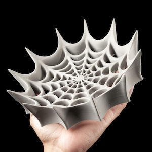 Op de afbeelding: Een grijze, spinnenwebvormige decoratieve kom met een gedetailleerd webontwerp. De kom heeft een reeks puntige randen en wordt in een hand gehouden tegen een zwarte achtergrond. De kom is een uniek, artistiek stuk.