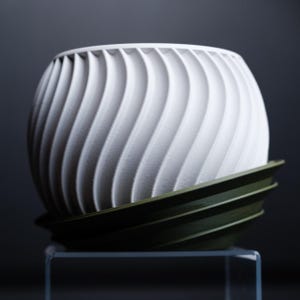 Op de afbeelding: Witte plantenbak met een draaiend, gestructureerd ontwerp. De plantenbak heeft een donkergroene, geribbelde basis.