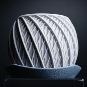 Può includere: Un vaso da fiori in ceramica bianca con un design geometrico intrecciato. Il vaso ha una base larga e rotonda e un'apertura stretta nella parte superiore.