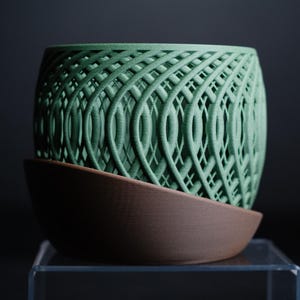 Può includere: Un vaso verde con un motivo intrecciato è posto su un piattino marrone. Il vaso ha una forma cilindrica con un intricato design a trama aperta. Il piattino ha una forma semplice e curva, che fornisce una base contrastante.