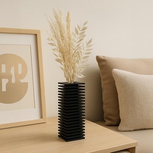 May include: A black, geometric vase with a layered design holds dried, beige-colored decorative grasses. The vase sits on a light-colored wooden surface, next to a framed artwork and a beige pillow.
