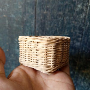 Puede incluir: Una pequeña cesta cuadrada tejida de mimbre natural. La cesta es de color marrón claro y tiene un diseño sencillo y rústico.