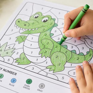 May include: A color-by-number coloring page featuring a cartoon alligator is being colored with a green crayon. The page has numbered sections to guide the coloring process. The text "COLOR BY NUMBER" is visible at the bottom of the page.