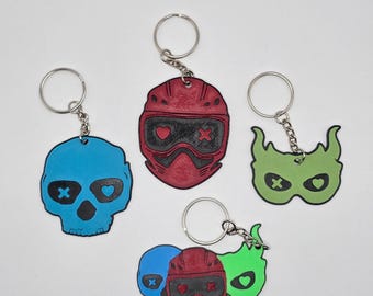 Pode incluir: Uma coleção de chaveiros com designs únicos. Os chaveiros incluem uma caveira azul, um capacete vermelho e uma máscara verde, cada um com detalhes de coração e cruz. Os chaveiros são presos a argolas prateadas.