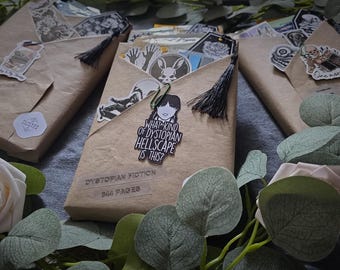 Pode incluir: Três pacotes de papel castanho amarrados com barbante preto e decorados com vários adesivos. O pacote do meio tem um adesivo que diz "Que tipo de inferno distópico é este?" e um adesivo de uma menina com um corte de cabelo preto.