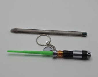 Pode incluir: Uma caneta prateada e um porta-chaves de sabre de luz são exibidos em fundo branco. A caneta tem uma ponta verde. O porta-chaves de sabre de luz tem uma lâmina verde, cabo prateado e preto e um porta-chaves.