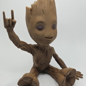 Puede incluir: Una figura marrón que parece un árbol con una superficie texturizada similar a la corteza. La figura está sentada con un brazo levantado, haciendo un gesto con la mano con el dedo índice y el meñique extendidos. La parte superior de la cabeza está abierta, revelando una pequeña planta.
