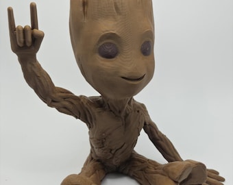 Pode incluir: Uma figura castanha semelhante a uma árvore com uma superfície texturizada que lembra casca. A figura está numa posição sentada com um braço levantado, fazendo um gesto com a mão com o dedo indicador e o dedo mindinho estendidos. O topo da cabeça está aberto, revelando uma pequena planta.