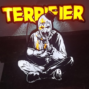 Może przedstawiać: Znak z napisem "TERRIFIER" w jaskrawożółtych literach. Poniżej znak czarno-biała ilustracja siedzącej postaci podobnej do klauna z palcem na ustach. Postać ma białą twarz z żółtymi akcentami.