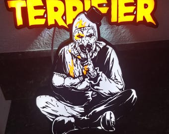 Pode incluir: Uma placa com a palavra "TERRIFIER" em letras amarelo brilhante. Abaixo da placa, uma ilustração em preto e branco de uma figura semelhante a um palhaço sentado com um dedo nos lábios. A figura tem um rosto branco com detalhes amarelos.