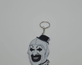 Pode incluir: Um chaveiro preto e branco com uma imagem estilizada do rosto de um palhaço com uma cartola. O palhaço tem uma boca larga e sorridente e maquiagem preta nos olhos. O chaveiro é preso a um anel de metal prateado.