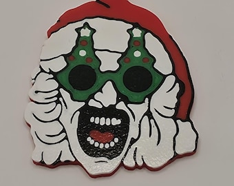 Pode incluir: Um porta-chaves de borracha com uma personagem de rosto branco e boca aberta, usando um gorro de Pai Natal vermelho e óculos verdes em forma de árvores de Natal. O design inclui contornos pretos e um anel de metal prateado com corrente.