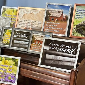 May include: A collection of framed art prints with various images and text. The prints feature floral arrangements, landscapes, and inspirational quotes. Frames are in various colors and styles, displayed on a wooden surface.