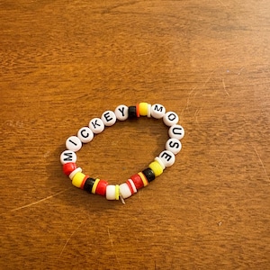 Peut inclure: Un bracelet de perles épelant "MICKEY MOUSE" avec des perles de lettres blanches. Le bracelet comporte des perles d'accentuation rouges, noires, jaunes et blanches. Le bracelet est sur une surface en bois.