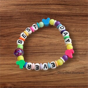 May include: A colorful beaded bracelet with the words "BEST DAY EVER" spelled out in white letter beads. The bracelet features various colored beads, including blue Mickey Mouse-shaped beads and pink heart-shaped beads. The bracelet is on a wooden surface.
