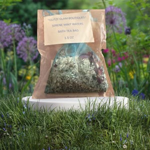 Puede incluir: Una bolsa de té de baño transparente, triangular, llena de hierbas y flores secas, exhibida sobre un pedestal blanco. La bolsa está etiquetada como "Serene Mint Waters" y "1.5 OZ". El embalaje incluye un respaldo de papel marrón y una cinta azul.