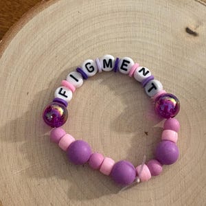 May include: A beaded bracelet with the word "FIGMENT" spelled out in white letter beads. The bracelet features a mix of pink, purple, and iridescent beads. The bracelet is displayed on a wooden surface.