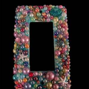 Könnte beinhalten: Eine rechteckige Lichtschalterabdeckung, verziert mit einer Vielzahl bunter Perlen und kleinen maritimen Charms. Der Rahmen ist hellblau lackiert und zeigt Perlen, Seesterne und ein Seepferdchen. Das Gesamtdesign ist ein lebendiges, vom Ozean inspiriertes Thema.
