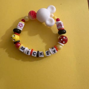 Op de afbeelding: Een kleurrijke kralenarmband met de naam "MICKEY" in letterkralen. De armband heeft een witte Mickey Mouse-hoofdbedeltje en diverse kralen in rood, geel, zwart en wit. De armband ligt op een gele achtergrond.