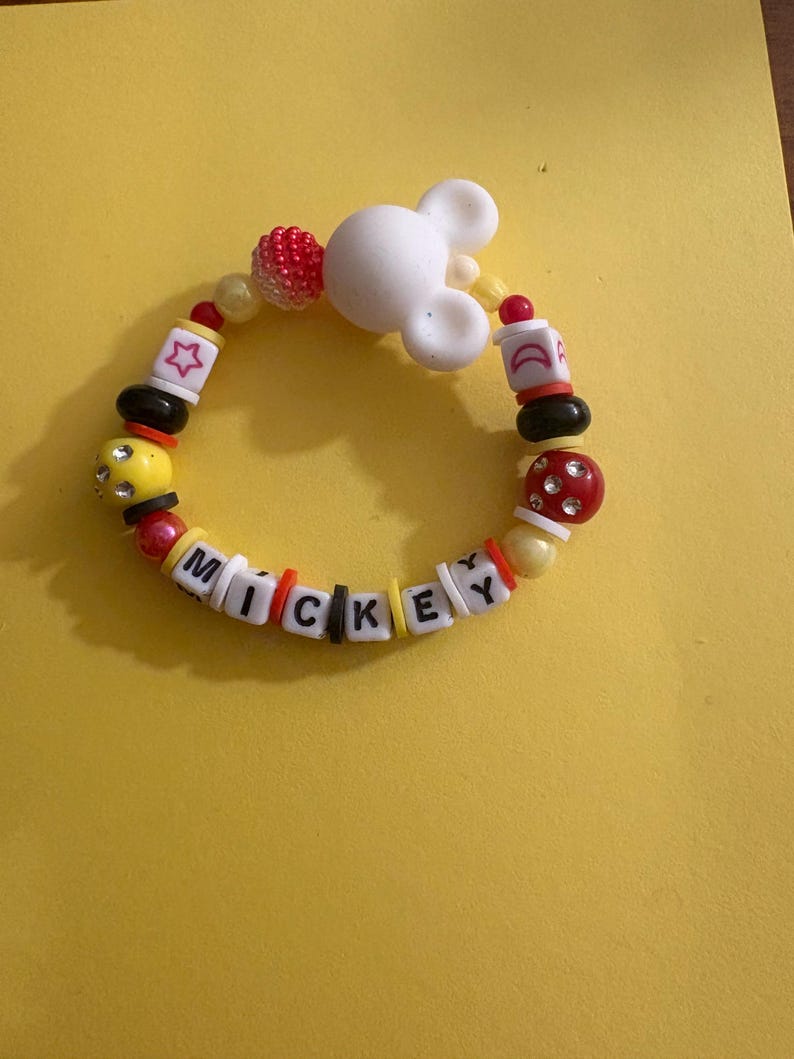 Op de afbeelding: Een kleurrijke kralenarmband met de naam "MICKEY" in letterkralen. De armband heeft een witte Mickey Mouse-hoofdbedeltje en diverse kralen in rood, geel, zwart en wit. De armband ligt op een gele achtergrond.