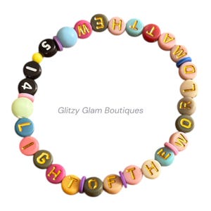 May include: A colorful beaded bracelet spelling out "LIGHT OF THE WORLD" with gold lettering. The bracelet features a variety of pastel-colored beads and round letter beads. The brand name "Glitzy Glam Boutiques" is printed on the image.