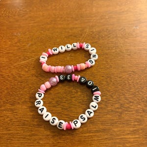 May include: Two handmade beaded bracelets. One bracelet is pink and white with the words "I WILL SING". The other bracelet is pink, black, and white with the words "PRAISE PSALM 104".